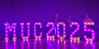 Violet illuminated letters stand out against a purple background. Behind them are Hendrik Sellmann, Frauke Mörike, and Jana-Sophie Effert leaning against the illuminated letters.