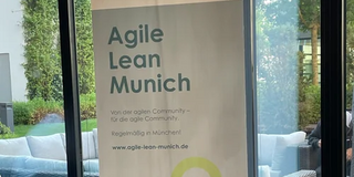 Vor einem Fenster steht ein Schild mit dem Text: "Agile Lean Munich. Von der agilen Community - für die agile Community. Regelmäßig in München! www.agile-lean-munich.de"