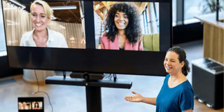 Hybride Arbeitstreffen Eine Person, welche präsent im Raum steht, stellt zwei weitere Personen vor, welche auf einem Bildschirm zu sehen sind.