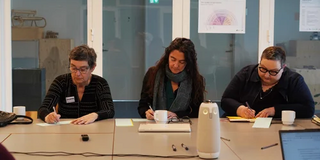 Drei Teilnehmende des Symposiums sitzen an einem Konferenztisch und schreiben.