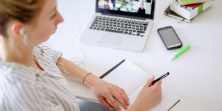Digitales Lernen Am Schreibtisch sitzende Studentin schaut ein Lernvideo, daneben liegen Bücher und Stifte.